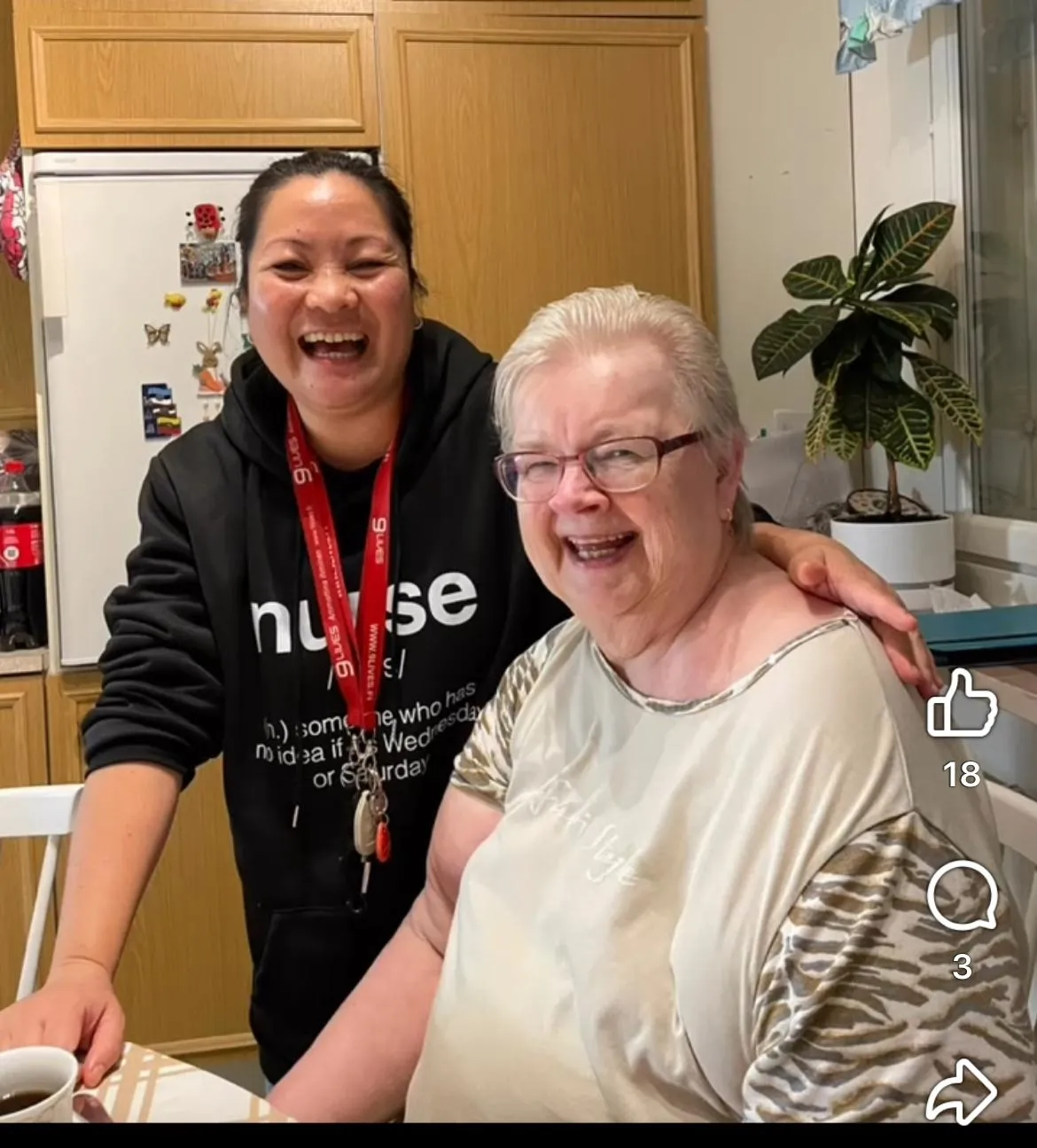 Kaksi iloista naista hymyilemässä ja halaamassa keittiössä, taustalla jääkaappi ja kasvi ikkunan vieressä.